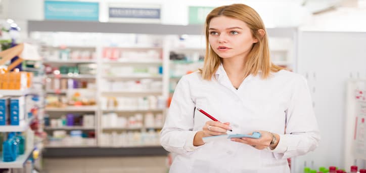 A woman in a white coat, writing on a clipboard while looking at a generic medication box. 